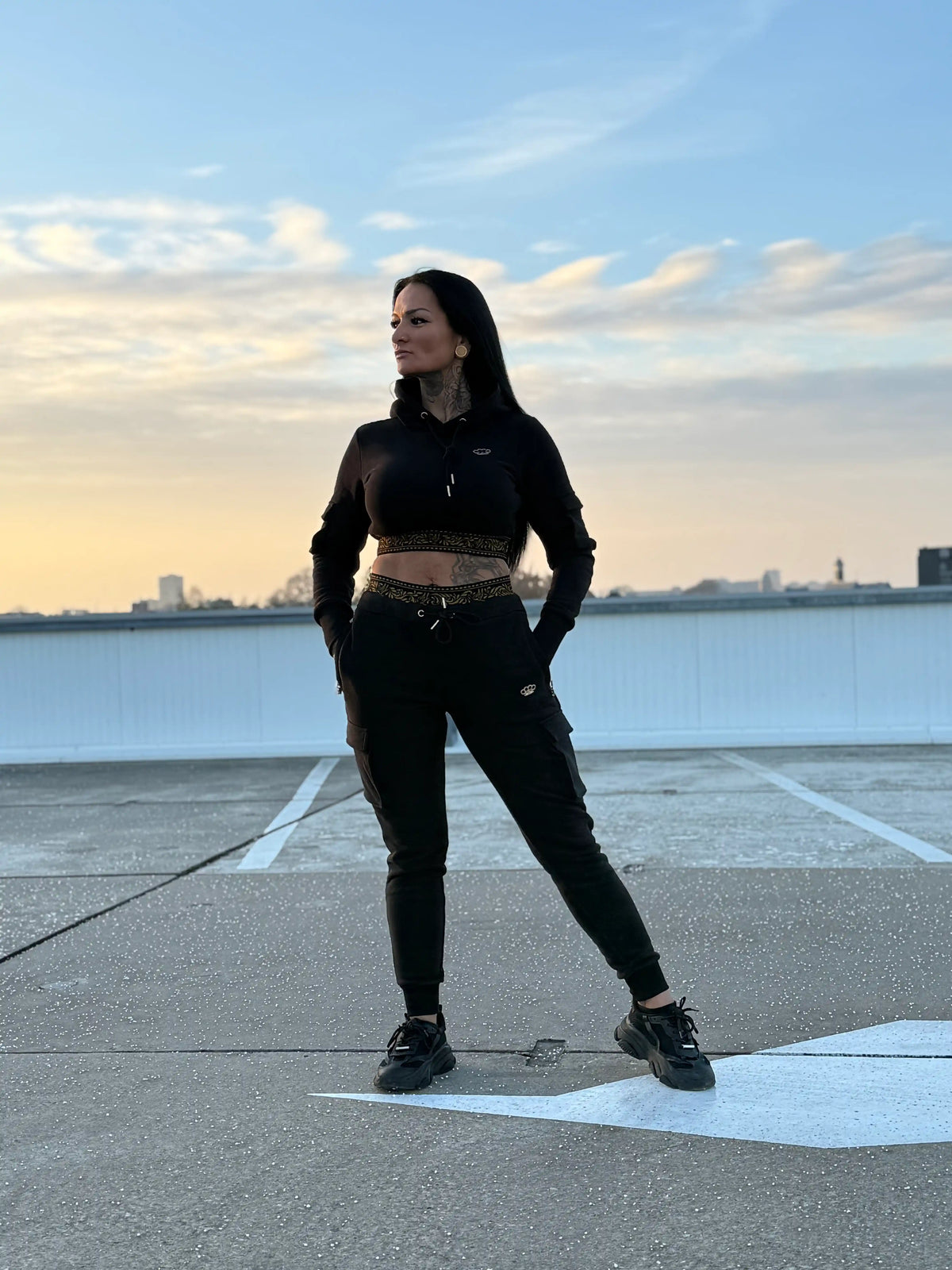 The full black MVL Meander women's tracksuit with gold bands, standing on a rooftop under a blue sky