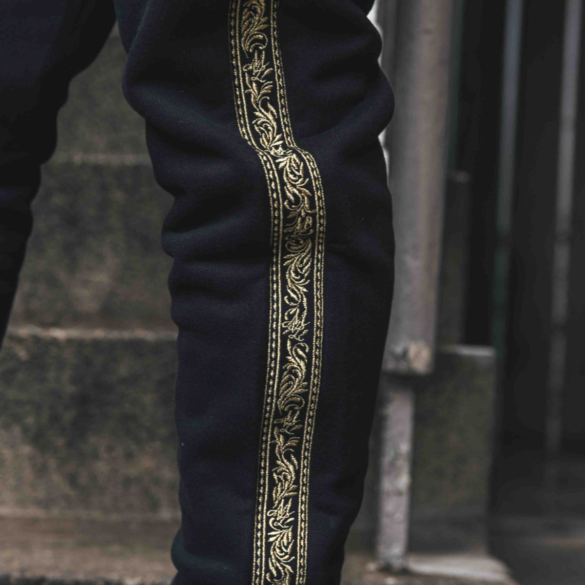 Close-up van de gouden Meander embroidery band langs de pijp van de zwarte MVL joggingbroek